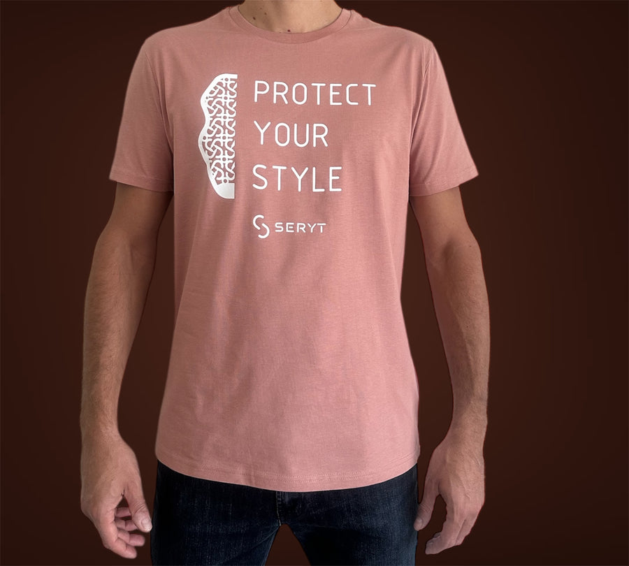 A man wearing a dusty rose pink t-shirt with a white graphic print that reads 'PROTECT YOUR STYLE' and features the SERYT brand logo. The background is a solid dark brown studio backdrop.